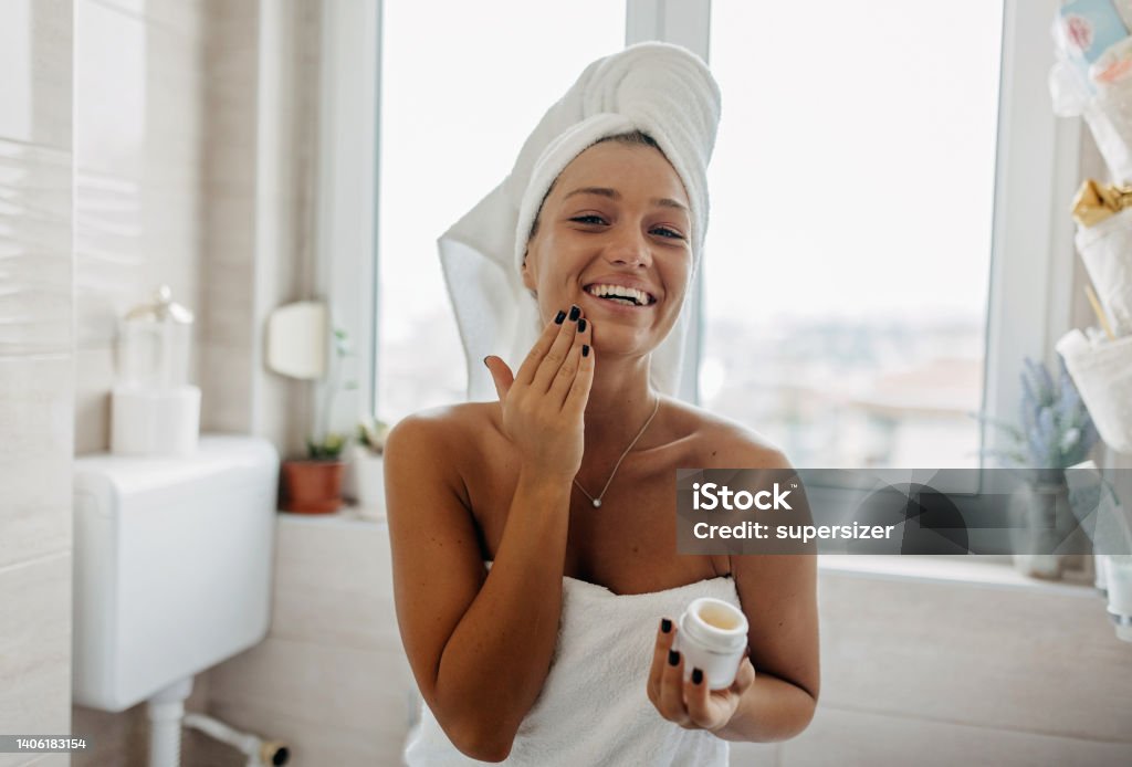 My face feels so soft and clean A young Caucasian woman is looking at the camera with a big smile on her face, while applying a face cream and wearing her hair up in a towel. Women Stock Photo