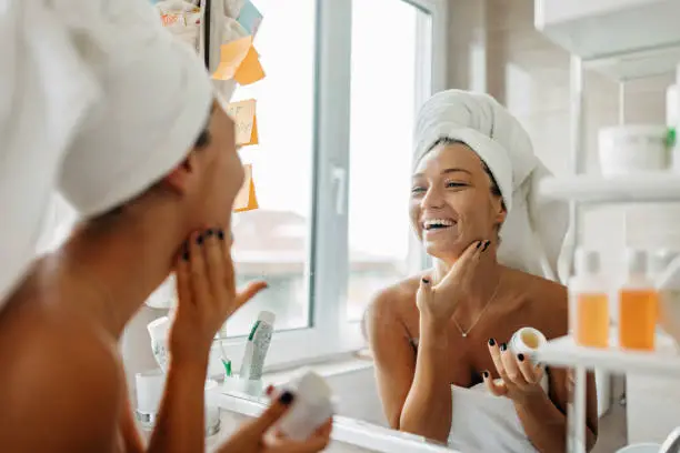 A beautiful young Caucasian woman is looking at the mirror with a big smile, while putting on her skin care products.