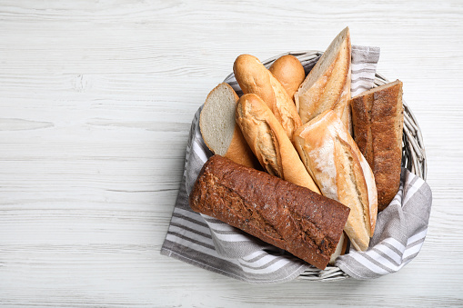 Different tasty baguettes in basket on white wooden table, top view. Space for text