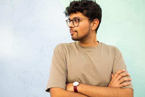 Portrait of a young handsome Indian man looking away . He is leaning on light blue wall
