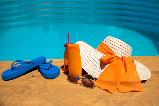 Sun lotion, Flip flops, sunglasses, swimsuit and towel on the wooden poolside deck. Summer accessories on the pool. Blu and orange color Spa Holiday background. Copy space