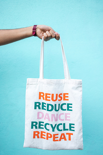 Hand holds a fabric shopping bag on the light blue wall. Bag has a message Reuse, Reduce, Dance, Recycle, Repet