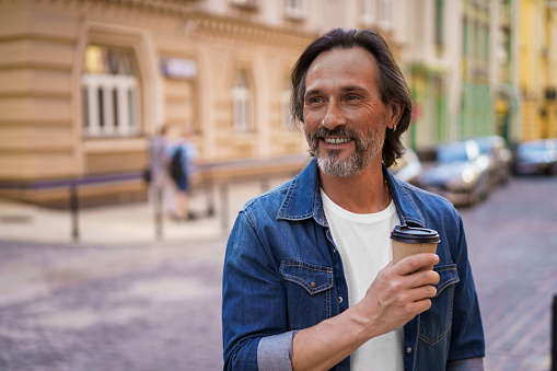 Attractive mature freelancer man smiling looking sideways holding disposable paper cup with hot coffee standing at old town. Handsome middle aged man with take away cup of coffee at the lunch break.