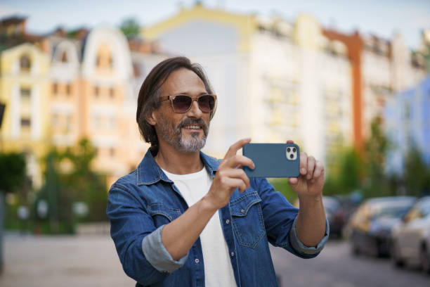 istock Happy middle aged grey bearded man taking pictures using smartphone camera in old town wearing casual. Happy senior man on street of european city with mobile phone. Travel concept 1406201906