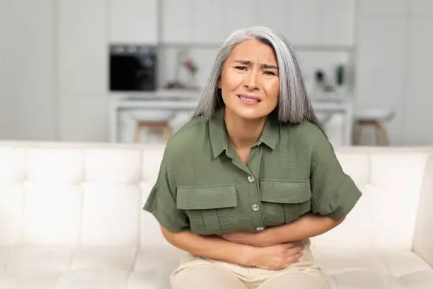 Concerned mature 50s woman with gray-hair suffering from stomach-ache sitting on sofa, middle-aged female holding tummy, undergoing belly pain and discomfort, suffer from menstruation