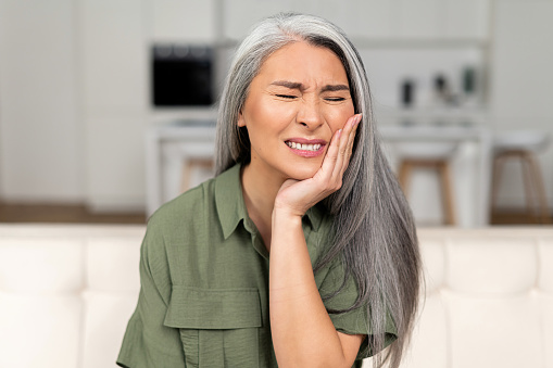Troubled mature 50s woman with gray-hair feels strong toothache, holding cheek and suffering from strong dental pain sitting on sofa, middle-aged female undergoing tooth pain, enamel sensitivity