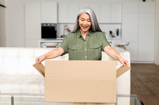Excited positive middle-aged Asian woman opening cardboard box, mid-age multiracial customer received an online order by postal shipment, astonished gray-haired lady unpacking parcel at home