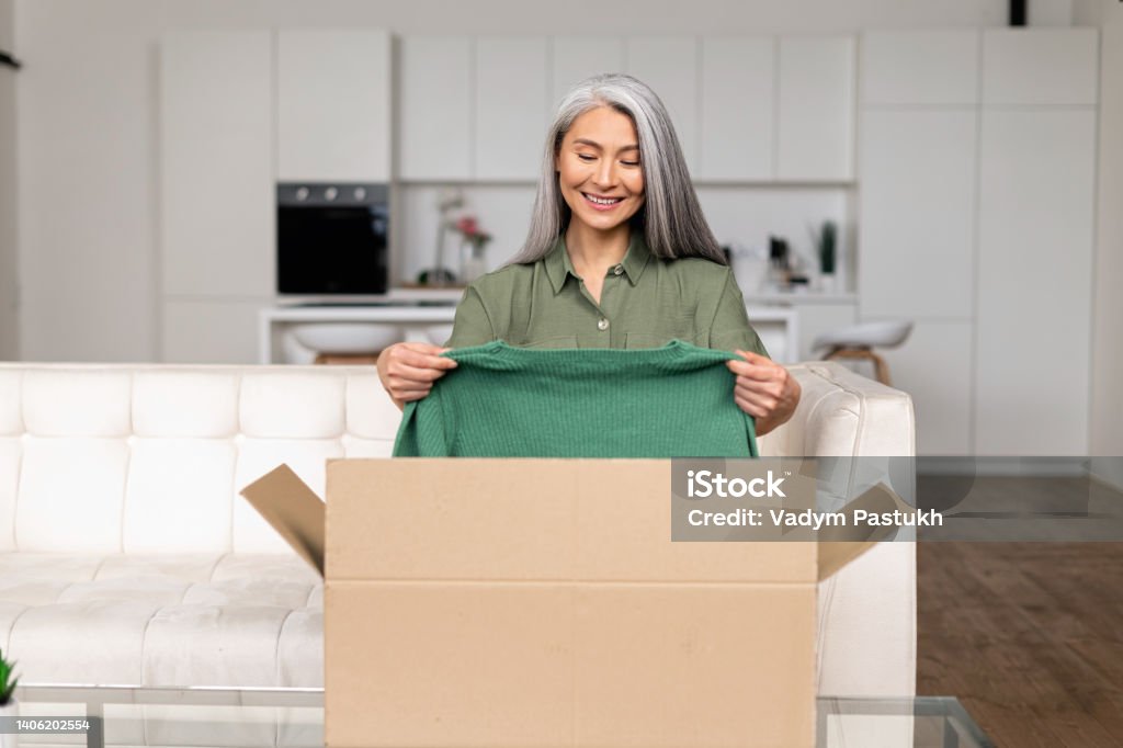 Positive mature asian woman standing with carton box and taking out new clothes Positive mature asian woman standing with carton box and taking out new clothes, while feeling curious about ordered item from online store. Smiling client satisfied with fast courier delivery service 45-49 Years Stock Photo
