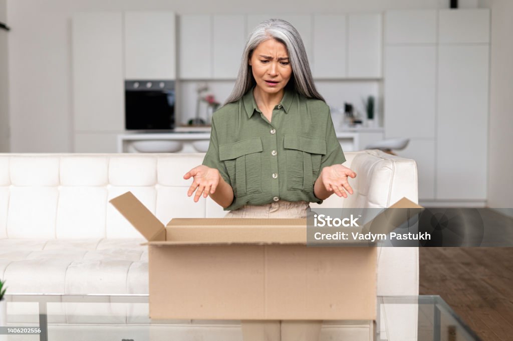 Disappointed middle-aged Asian lady upset with an online purchase Disappointed middle-aged Asian lady upset with an online purchase. Gray-haired woman looking inside cardboard shipping box and feels unhappy and angry with wrong order Mistake Stock Photo