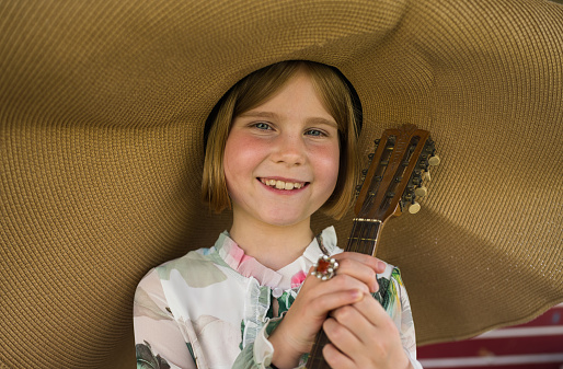 Portrait of a little girl. She wears a large beret hat. It's summer and she's about to perform a little garden concert with her mandolin.