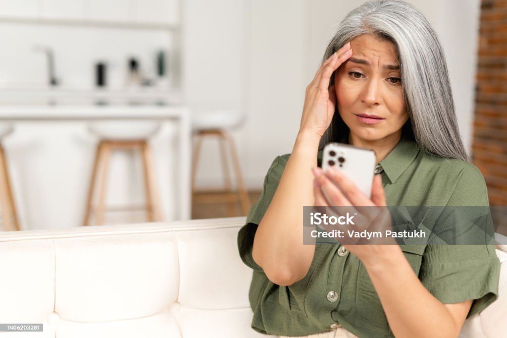 Frustrated unhappy gray-haired mature woman sitting on sofa, holding smartphone, senior female has not enough money for online transaction Frustrated unhappy gray-haired mature woman sitting on sofa, holding smartphone, senior female has not enough money for online transaction, financial problem Social Media Stock Photo