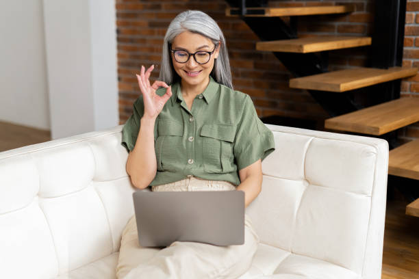 istock Modern mature female enjoy leisure weekend at home, browsing wireless internet on computer gadget and showing ok gesture. Happy elderly woman sit relax on couch in living room, work on laptop 1406203293