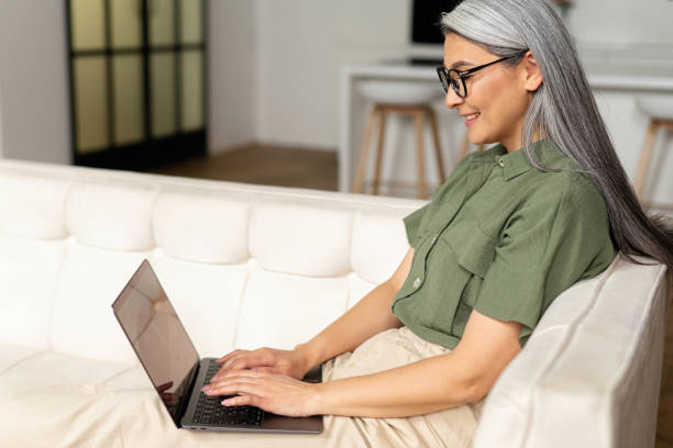 istock Smiling lady sitting at the cozy sofa and looking at the camera. Web browsing, watching TV series, shopping in network concept. Asian mature woman spending home leisure 1406203327