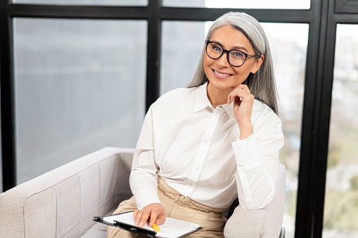 Business portrait of a confident beautiful senior female businesswoman in glasses looking straight at the camera, holding clipboard. Elder smiling woman is a professional lawyer or psychologist