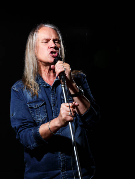 elderly man with long gray hair in blue denim shirt sings into a microphone on stage on black background