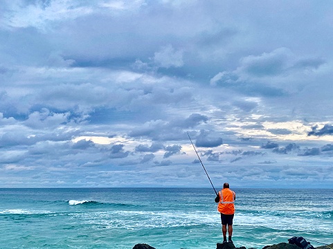 Horizontal seascape of fisherman fishing off waters edge promenade rock wall into turquoise wild stormy ocean under a grey cloudscape moody sky at Brunswick Heads near Byron Bay NSW Australia