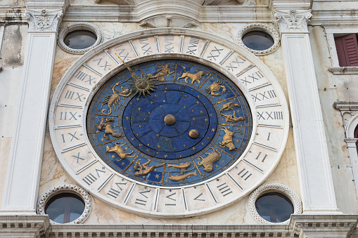 Venice, Italy - September 24, 2021: Clock face detail of St. Mark's Clocktower in Venice. The Clock Tower in Venice is an early Renaissance building on the north side of the Piazza San Marco. It comprises a tower, which contains the clock. A fixed circle of marble is engraved with the 24 hours of the day in Roman numerals. A golden pointer with an image of the sun moves round this circle and indicates the hour of the day. Within the marble circle beneath the sun pointer are the signs of the zodiac in gold which revolve slightly more slowly than the pointer to show the position of the sun in the zodiac. In the middle of the clockface is the earth (in the centre) and the moon, which revolves to show its phases, surrounded by stars which are fixed in position.