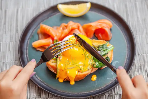 Photo of Woman hands cut by fork and knife fresh eggs Benedict with smoked salmon on plate in restaurant. Gourmet hotel food with baked toast bread, healthy snack. Delicious lunch in cafe