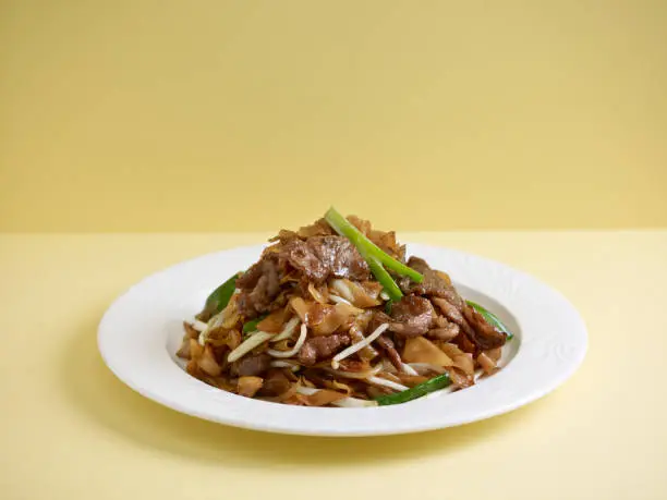 Photo of wok-fried Hor Fun with sliced beef served in a dish isolated on wooden board side view on grey background