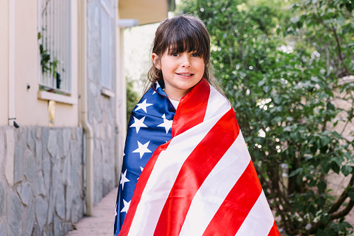 Black-haired girl covered with a large American flag, in the garden of her house. Concept of celebration, independence day, United States of America, 4th of July, patriotism and American pride.