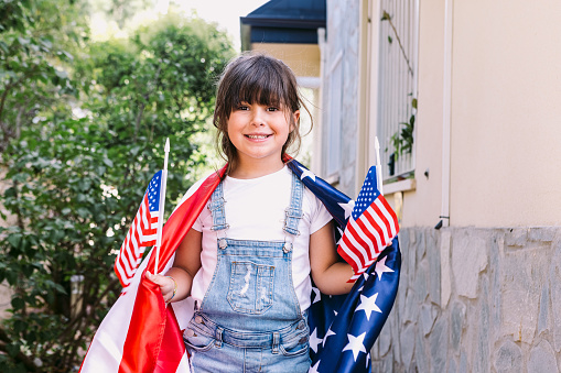 Little black-haired girl covered with big USA flag, holding little American flags in her home garden. Concept of celebration, independence day, 4th of July, patriotism, holiday and American pride.