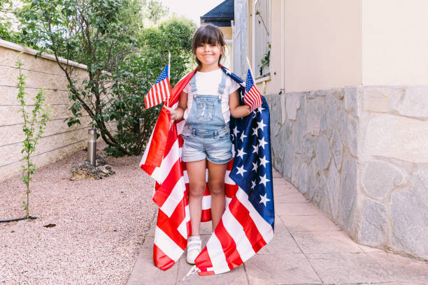 Little black-haired girl covered with big USA flag, holding little American flags in her home garden. Concept of celebration, independence day, 4th of July, patriotism, holiday and American pride. stock photo