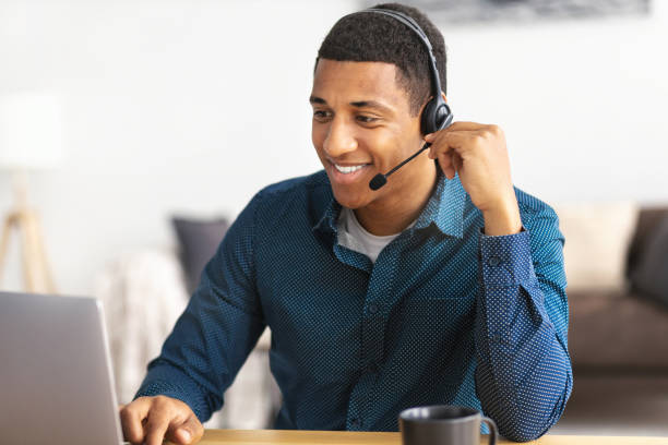 giovane afroamericano operatore di call center o receptionist seduto sul posto di lavoro in un ufficio moderno che consulta un cliente, usa un auricolare, sorride amichevole - sales agent foto e immagini stock