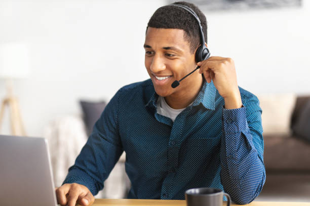 african american young man customer support call center operator or receptionist sitting at the workplace in a modern office consulting a client, uses a headset, smiles friendly - receptionist customer service customer service representative imagens e fotografias de stock