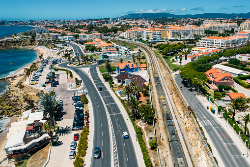 Aerial drone view of train passing near the Marginal Avenue and coastline with Parade district in Greater Lisbon, Portugal looking west towards Cascais and Sintra hills