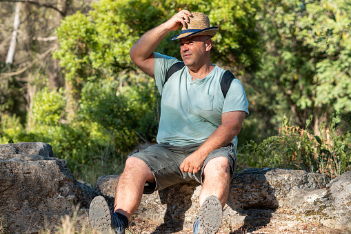 Overweight sweaty man, sitting on a rock in the forest, putting on a straw hat and a backpack on his back.