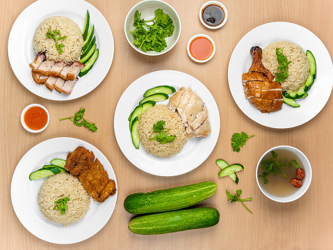 Chicken Cutlet and Roast Pork Rice, Roast Pork, Roasted Chicken, Roast Pork, Steamed Chicken with raw cucumber, Coriander, salad, sauce and soup served in a dish isolated on wooden background top view