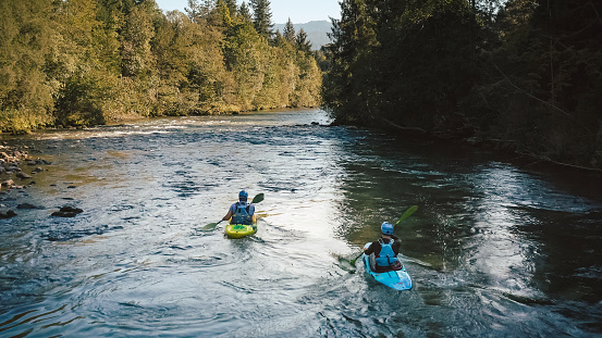 Two men in kayaks, paddling over a mountain river, enjoying the view of beautiful green forest coast, aerial tracking shot.