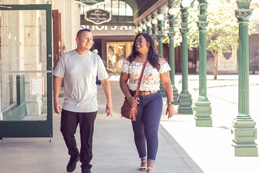 A hispanic man and African American woman walk together down a covered sidewalk in front of local shops.