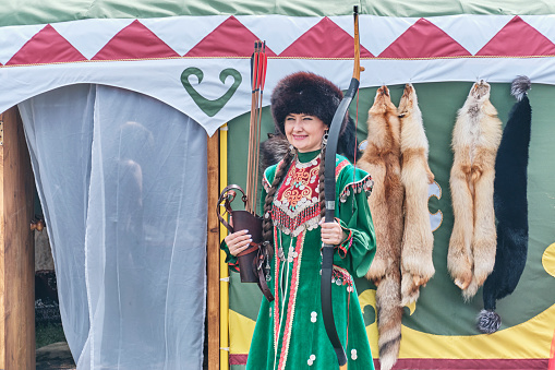 Yekaterinburg, Russia - June 25, 2022: Portrait of beautiful Asian woman in traditional festive Bashkir clothes, fur hat, guarding entrance to yurt with bow and arrow at Sabantuy folk festival.