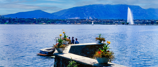 Geneva, Switzerland - July 10 , 2001 :  Geneva, Switzerland - July 10 , 2001 A few people standing by a motor boat with a sailboat and the shore with buildings and hills  in the rear.