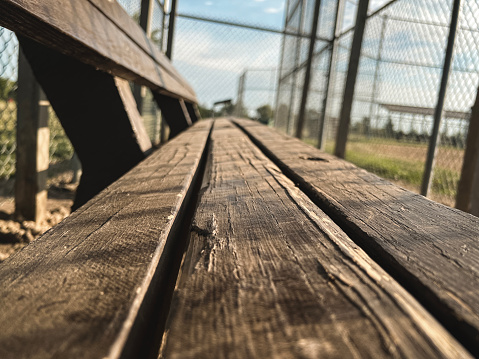 Worn brown wooden bench situated within a baseball or softball dugout. Sports field fencing in the background. Low-angle views.