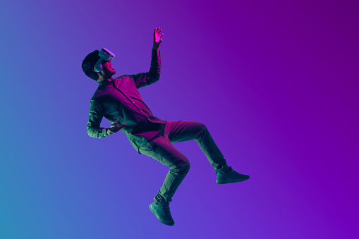 Young Asian man wearing VR headset enjoy playing video game and levitating in the air on futuristic purple cyberpunk neon light banner background. Metaverse technology concept.