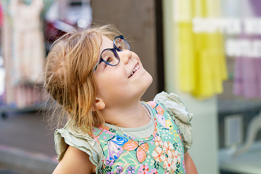 Portrait of a cute preschool girl with eye glasses outdoors. Happy funny child wearing new blue glasses. Sunny summer day in the city