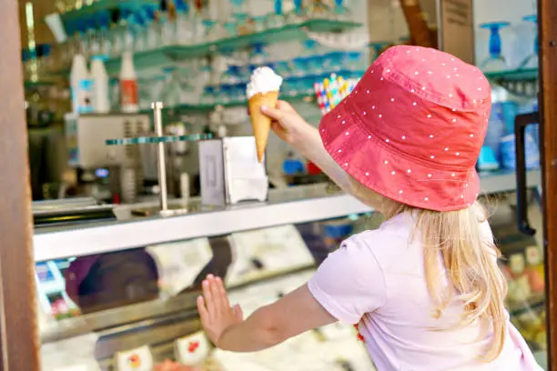 Photo of Cute little toddler girl choosing and buying ice cream in outdoor stand cafe. Happy preschool child with glasses looking at different sorts of icecream. Sweet summer dessert