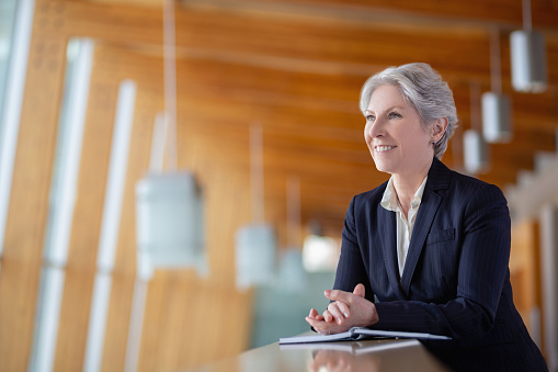 Portrait of smiling happy senior Caucasian businesswoman leaning on railing at office balcony with hands clasped