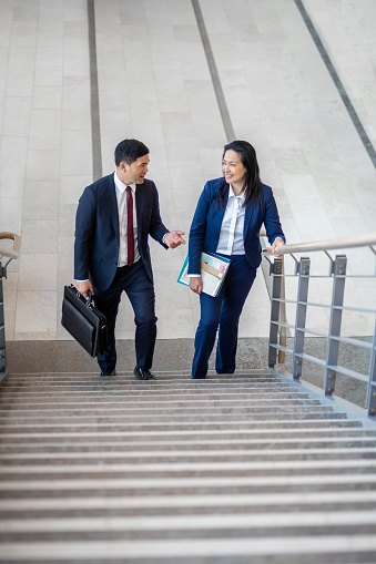 Pacific Islander ethnicity businessman with mature Asian businesswoman with briefcase climbing a staircase in office lobby