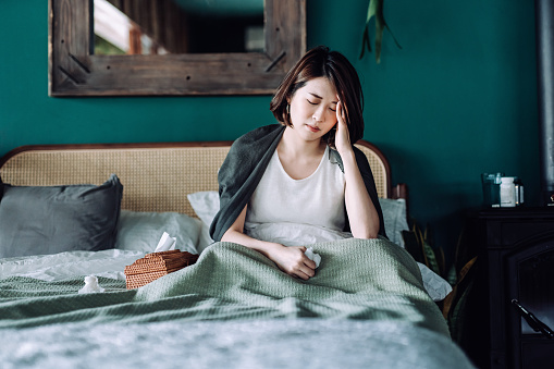 Young Asian woman staying at home and lying in bed, feeling sick with her hand on her head, with medicine bottle and a glass of water on the side table. Sickness, flu, fever, headache concept