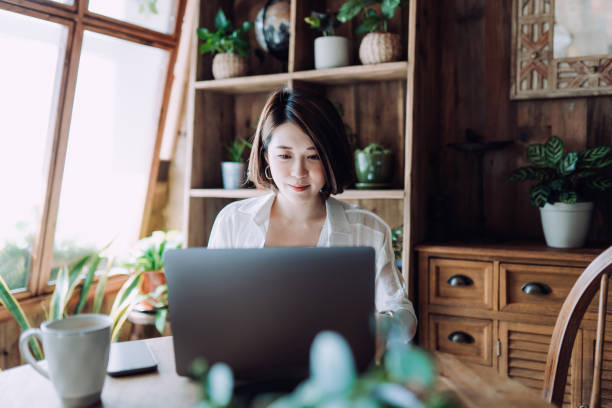 joven asiática independiente que trabaja en línea en una computadora portátil desde la oficina en casa. empresaria, emprendedora, bloguera, freelancer trabajando desde casa conceptos en un ambiente acogedor. estilo de vida y tecnología - trabajo freelance fotografías e imágenes de stock