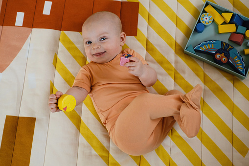 Infant on the floor of kids play room learning to crawl on the rug. Early development. Smiling Baby girl on sun indoors near toys on carpet. Movement and interaction. Cute six month old child at home