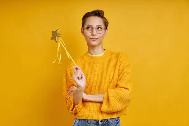 Playful woman holding magic wand and looking thoughtful while standing against yellow background