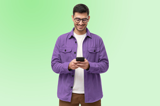 Portrait of handsome smiling hipster guy with stubble, in specs and stylish violet shirt standing against green gradient studio background, browsing social media on smartphone, chatting with friend
