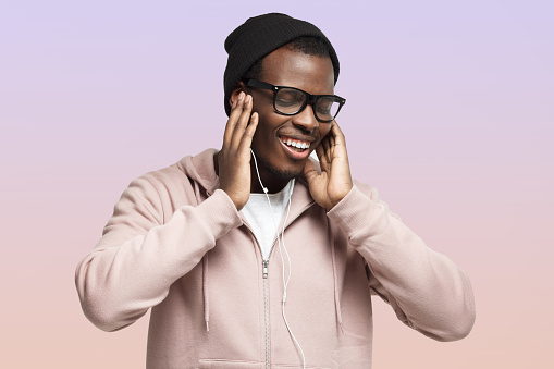 Portrait of relaxed happy smiling african american guy listening to music in wired earphones, enjoying favorite song, singing along with closed eyes, isolated on pink background