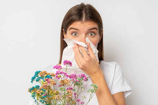 Close-up studio portrait of allergic girl in white clothes covering nose with tissue, having reaction to wild flowers in her hands. Seasonal pollen allergy concept