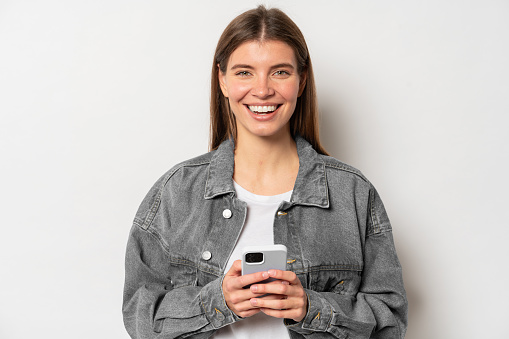 Cheerful young pretty woman with fair hair holding mobile phone laughing at memes, surfing internet, browsing social networks, web-pages, in denim jacket isolated over beige background