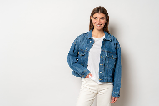 Pretty adorable youthful woman with fair hair dressed in white t-shirt, denim jacket and jeans, holding hand in pocket, smiling against blank studio wall with copy space. Fashion and trends concept